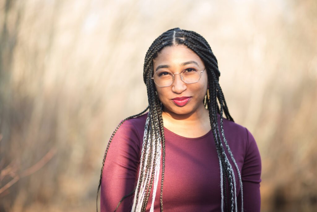 Picture of Dr. Dyette in a dark purple dress pictured from the waist up. She has gray and black braids in her hair and hexagon glasses and she is smiling.